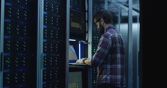 Adult bearded system administrator in glasses walks over to the server rack and opens the rack door in the server room and makes the hardware settings using a laptop.